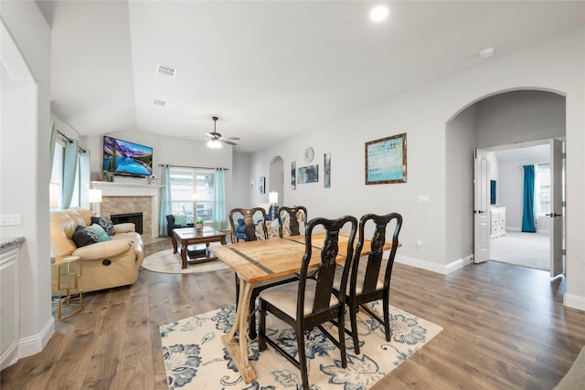 dining room featuring arched walkways, wood finished floors, a stone fireplace, ceiling fan, and baseboards