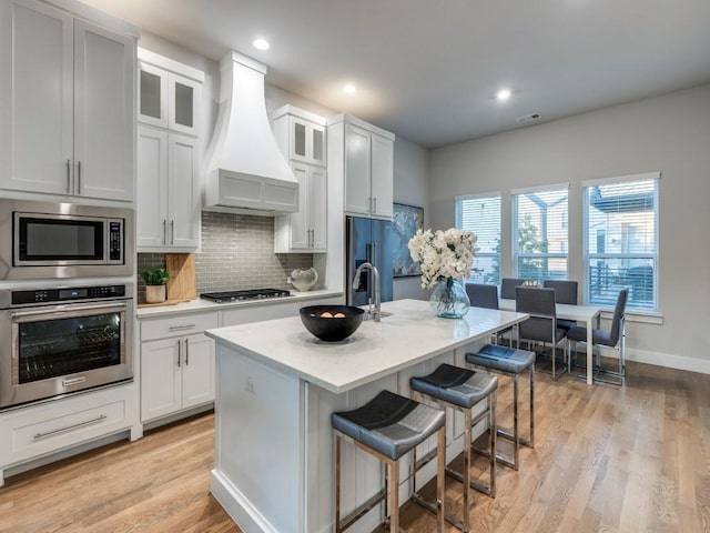kitchen with appliances with stainless steel finishes, premium range hood, a kitchen bar, tasteful backsplash, and light wood-style flooring