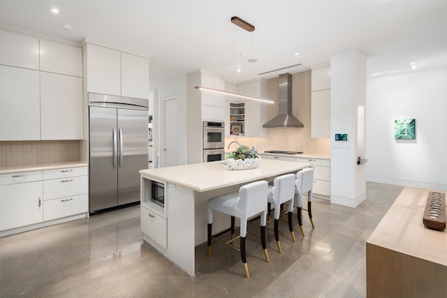 kitchen with built in appliances, wall chimney range hood, modern cabinets, a kitchen bar, and white cabinets
