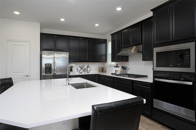 kitchen featuring stainless steel appliances, a sink, under cabinet range hood, dark cabinetry, and a kitchen island with sink