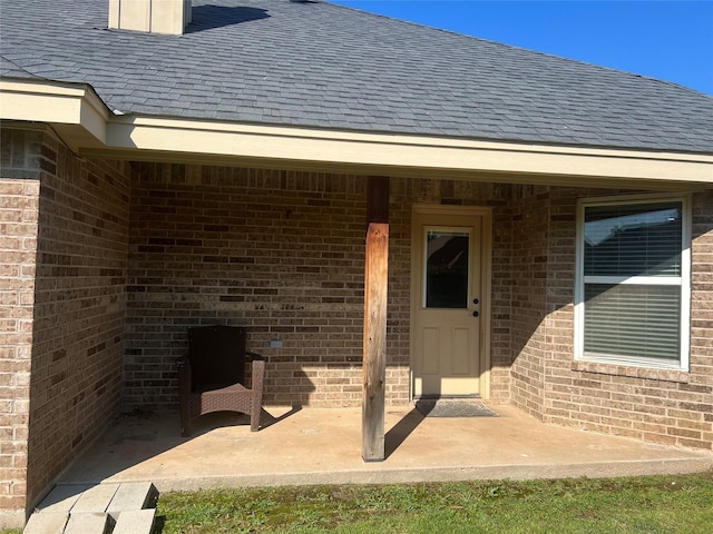 doorway to property featuring brick siding, a shingled roof, and a patio area