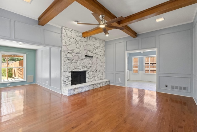 unfurnished living room featuring a decorative wall, ceiling fan, beamed ceiling, wood finished floors, and a stone fireplace