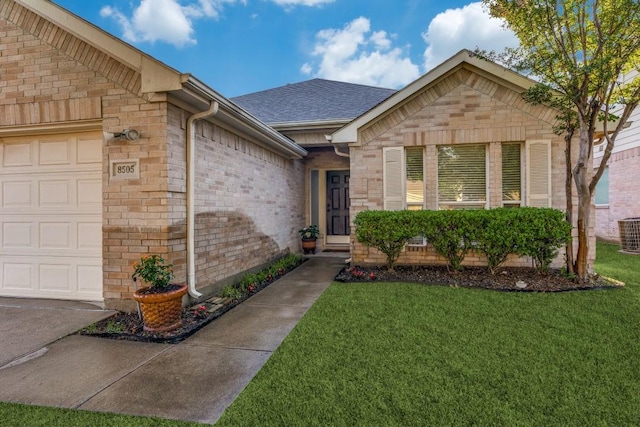 doorway to property with an attached garage, a yard, central AC, brick siding, and roof with shingles