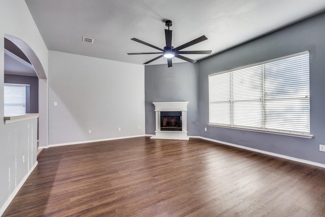 unfurnished living room featuring a ceiling fan, dark wood-style floors, a fireplace with raised hearth, baseboards, and arched walkways