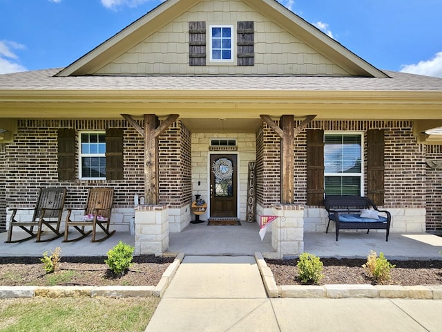 view of exterior entry featuring covered porch, brick siding, and roof with shingles