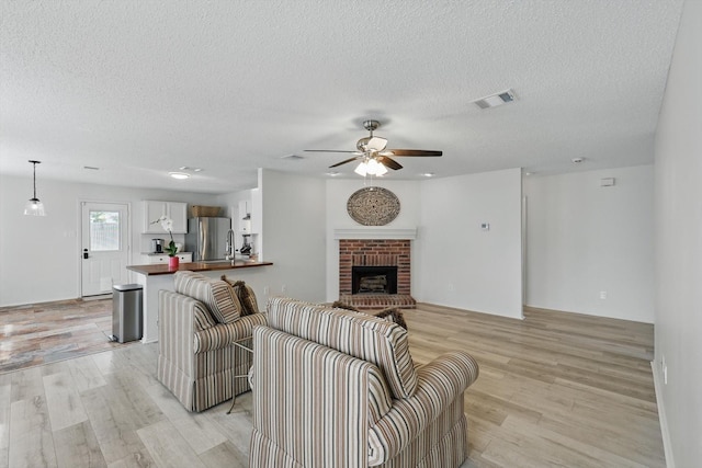 3110 Legend Dr, Mckinney, TX 75070 - living room featuring light wood-type flooring, a ceiling fan, a textured ceiling, and a brick fireplace