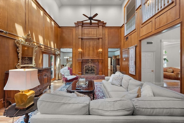 living room with a towering ceiling, wood finished floors, ceiling fan, a brick fireplace, and wooden walls