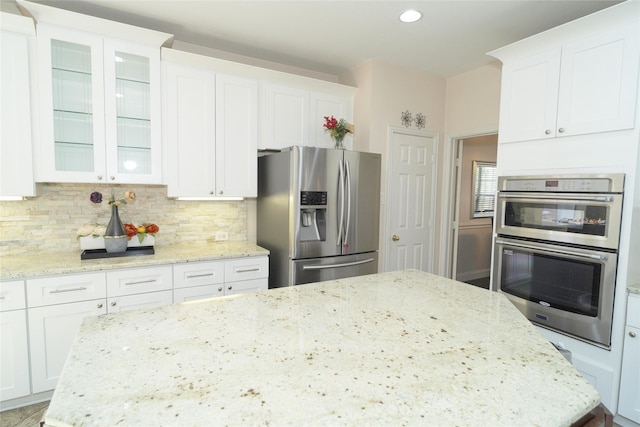 kitchen featuring appliances with stainless steel finishes, white cabinetry, backsplash, glass insert cabinets, and light stone countertops