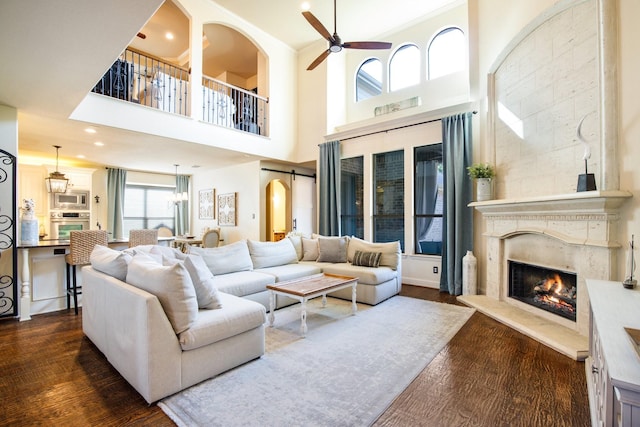 living room featuring a ceiling fan, a fireplace, dark wood-style floors, a high ceiling, and arched walkways