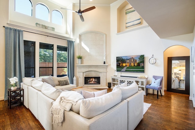 living area with a high ceiling, ceiling fan, arched walkways, wood finished floors, and a lit fireplace