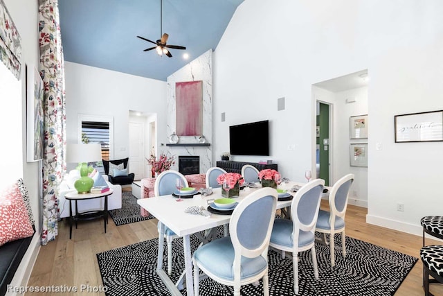 dining space featuring high vaulted ceiling, a fireplace, a ceiling fan, light wood-style floors, and baseboards