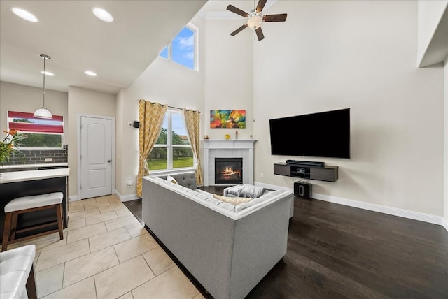 living room featuring a high ceiling, ceiling fan, a tiled fireplace, baseboards, and plenty of natural light