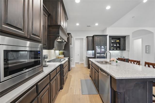 kitchen featuring stainless steel appliances, a sink, backsplash, a center island with sink, and dark brown cabinetry