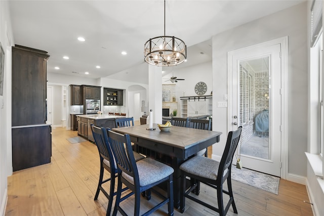 dining room with a ceiling fan, arched walkways, a chandelier, a large fireplace, and light wood-style floors