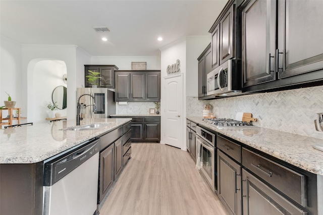kitchen with stainless steel appliances, crown molding, an island with sink, light stone countertops, and light wood-style floors