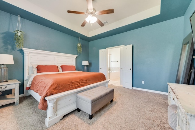 bedroom featuring a raised ceiling, light colored carpet, baseboards, ceiling fan, and ensuite bath