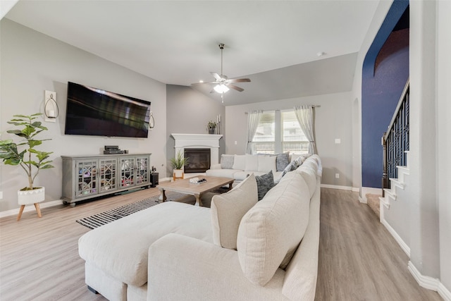 living room featuring light wood-type flooring, ceiling fan, baseboards, stairway, and a glass covered fireplace
