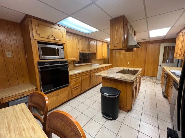 kitchen featuring black appliances, a drop ceiling, wood walls, brown cabinetry, and light tile patterned floors