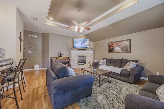 living area featuring a raised ceiling, ceiling fan, light wood-type flooring, baseboards, and a stone fireplace