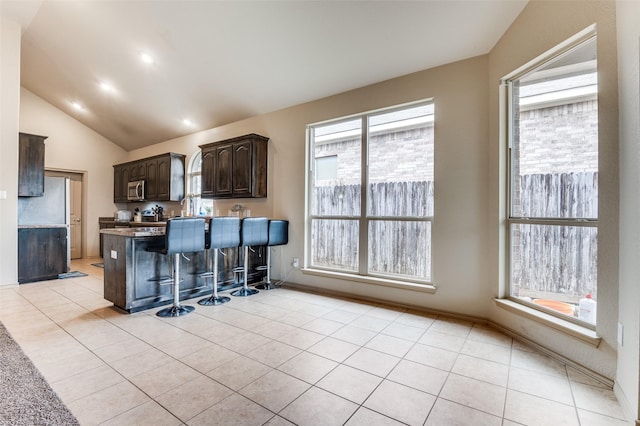 kitchen featuring dark brown cabinetry, light tile patterned flooring, a kitchen breakfast bar, vaulted ceiling, and stainless steel microwave