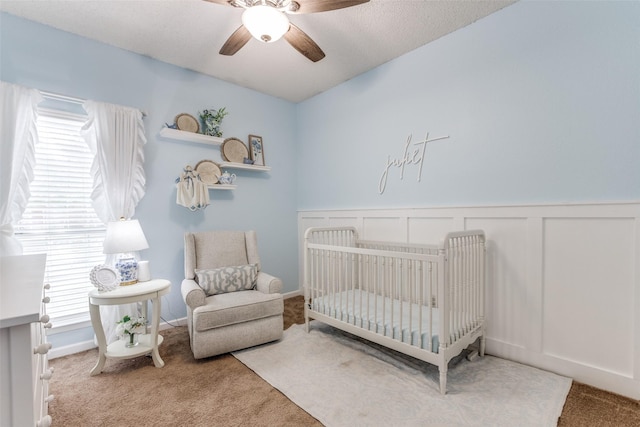 carpeted bedroom with a crib, a wainscoted wall, a ceiling fan, and a decorative wall