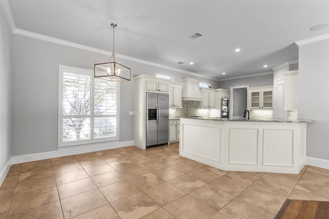 kitchen featuring stainless steel refrigerator with ice dispenser, custom range hood, a peninsula, light stone countertops, and decorative backsplash