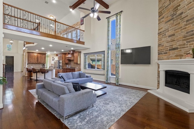 living room with a fireplace with raised hearth, dark wood-style floors, ceiling fan, a towering ceiling, and baseboards