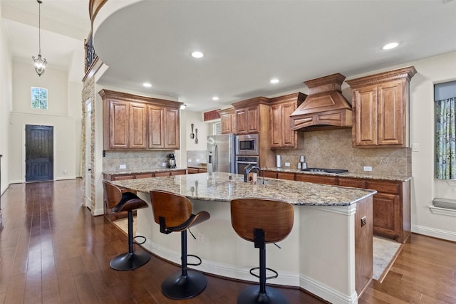kitchen featuring custom exhaust hood, appliances with stainless steel finishes, an island with sink, light stone countertops, and brown cabinetry