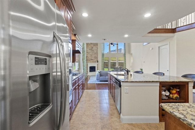 kitchen with stainless steel appliances, ceiling fan, open shelves, a large fireplace, and light stone countertops