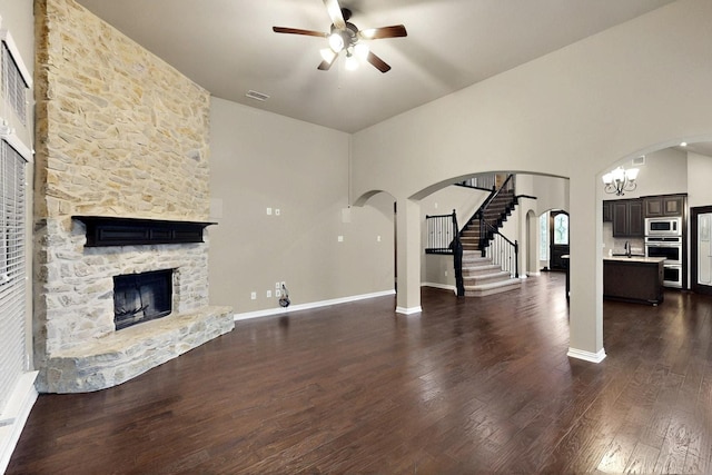 unfurnished living room featuring ceiling fan, arched walkways, a stone fireplace, dark wood-style flooring, and a towering ceiling