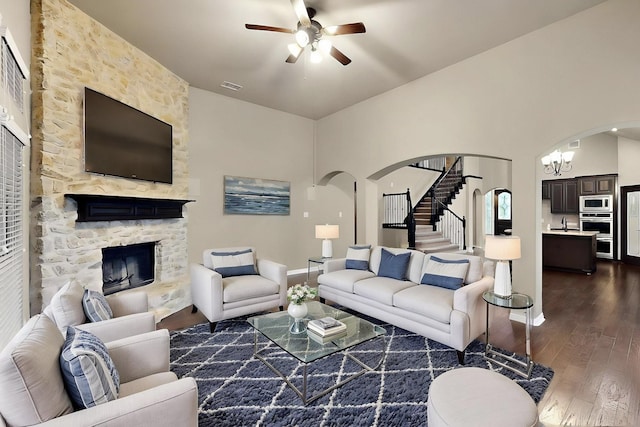 living area with arched walkways, a stone fireplace, a ceiling fan, dark wood-style flooring, and stairs