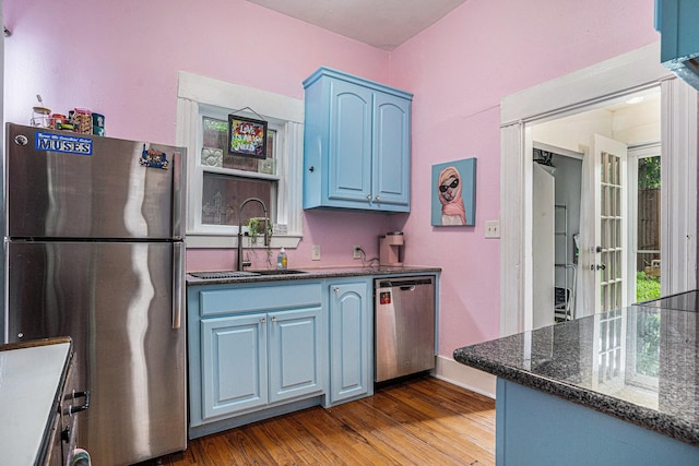 kitchen with appliances with stainless steel finishes, blue cabinetry, a sink, wood finished floors, and dark stone countertops