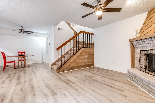 unfurnished living room with ceiling fan, wood finished floors, stairway, a brick fireplace, and a textured ceiling