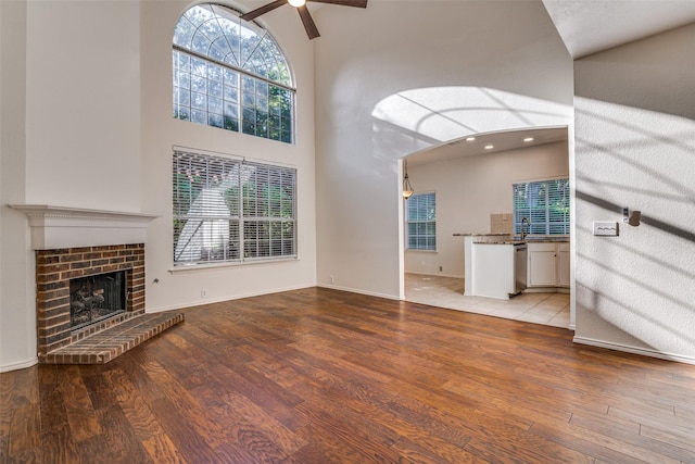 unfurnished living room featuring a high ceiling, wood finished floors, ceiling fan, a brick fireplace, and baseboards