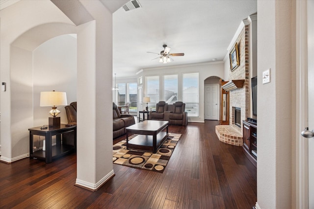 living area with ceiling fan, arched walkways, a fireplace, dark wood finished floors, and ornamental molding