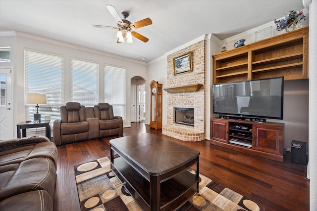 living area featuring arched walkways, ceiling fan, dark wood-style floors, ornamental molding, and a fireplace
