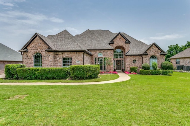 french country home with brick siding, a shingled roof, a front lawn, and central AC