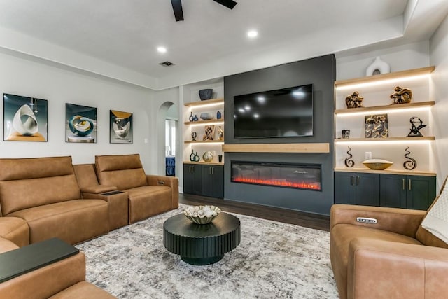 living room with arched walkways, a ceiling fan, dark wood finished floors, a glass covered fireplace, and built in shelves