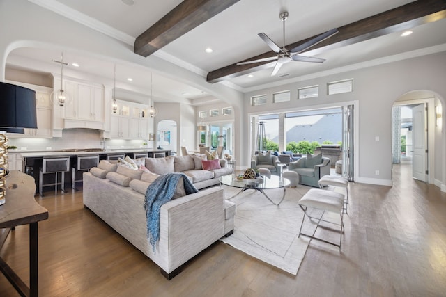 living area with arched walkways, a ceiling fan, beamed ceiling, wood finished floors, and crown molding