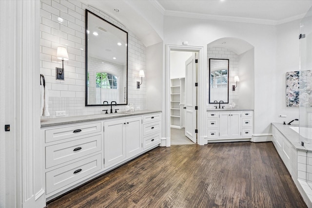 bathroom featuring wood finished floors, two vanities, decorative backsplash, ornamental molding, and a bath