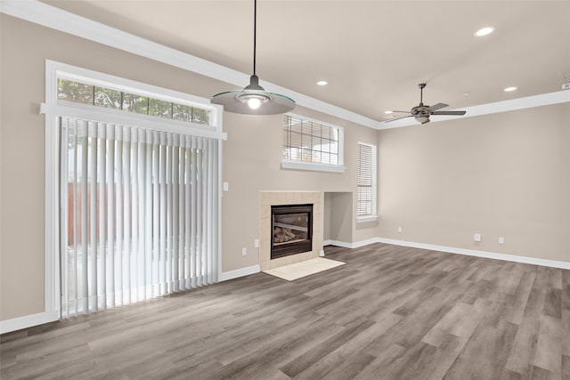 unfurnished living room featuring ceiling fan, healthy amount of natural light, wood finished floors, a tiled fireplace, and ornamental molding