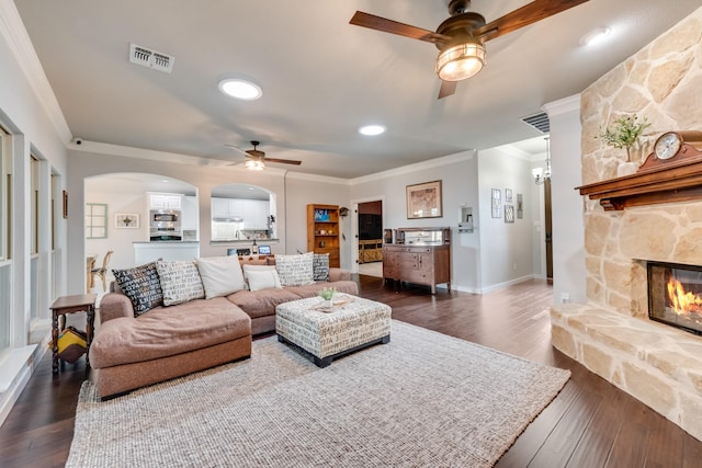 living room featuring ceiling fan, ornamental molding, dark wood-style flooring, a stone fireplace, and arched walkways