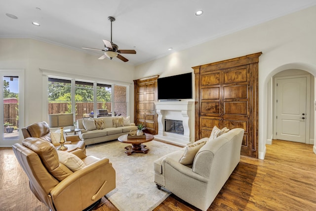 living area featuring arched walkways, wood finished floors, a ceiling fan, ornamental molding, and a fireplace with raised hearth