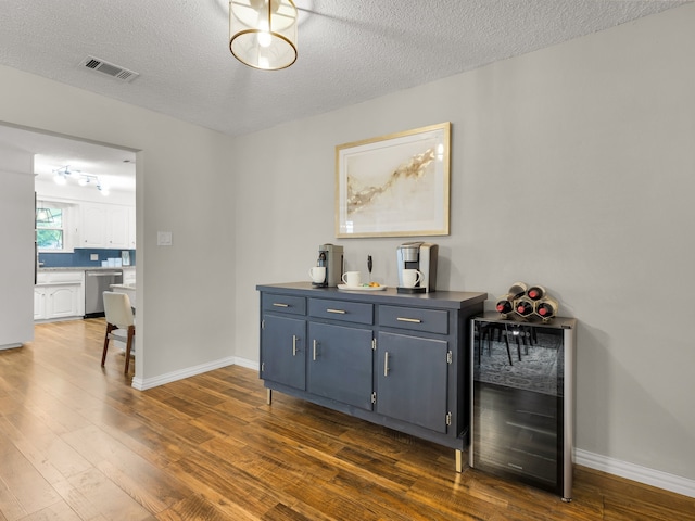 indoor bar featuring beverage cooler, stainless steel dishwasher, dark wood-type flooring, a textured ceiling, and baseboards