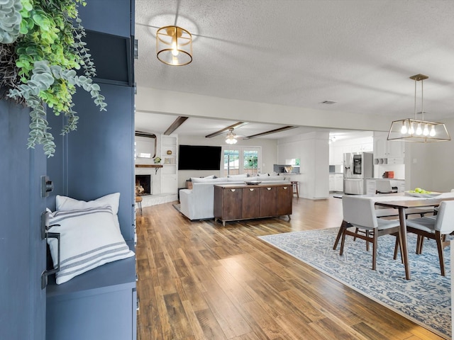 dining area featuring wood finished floors, beamed ceiling, a large fireplace, a textured ceiling, and ceiling fan