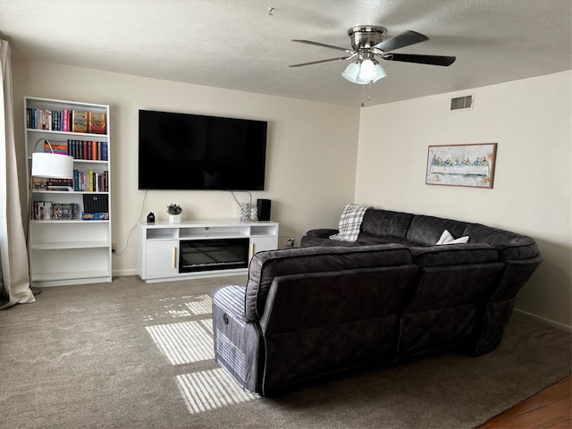 living room with a ceiling fan, a textured ceiling, carpet flooring, and a glass covered fireplace