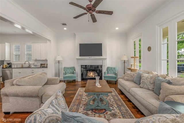 living area with a ceiling fan, dark wood finished floors, a warm lit fireplace, recessed lighting, and crown molding