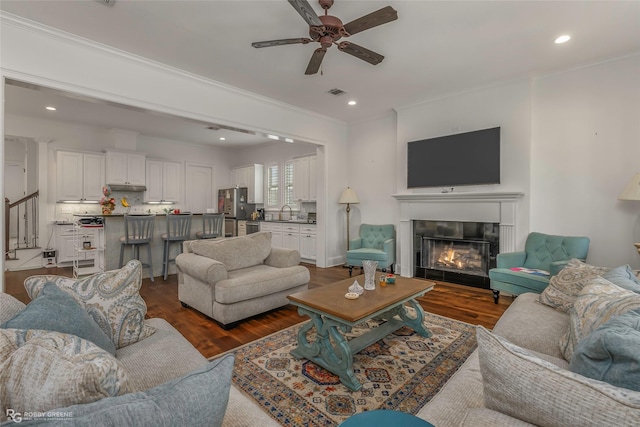 living area with ceiling fan, ornamental molding, a glass covered fireplace, dark wood-style flooring, and recessed lighting