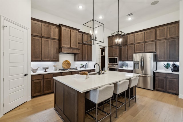 kitchen with appliances with stainless steel finishes, a chandelier, light wood-style flooring, backsplash, and dark brown cabinetry