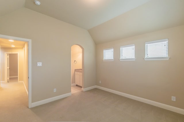 unfurnished bedroom with lofted ceiling, light carpet, arched walkways, baseboards, and connected bathroom
